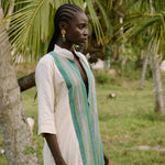 Woman in a long, light-colored dress with a green and white striped scarf standing in a natural setting with trees.