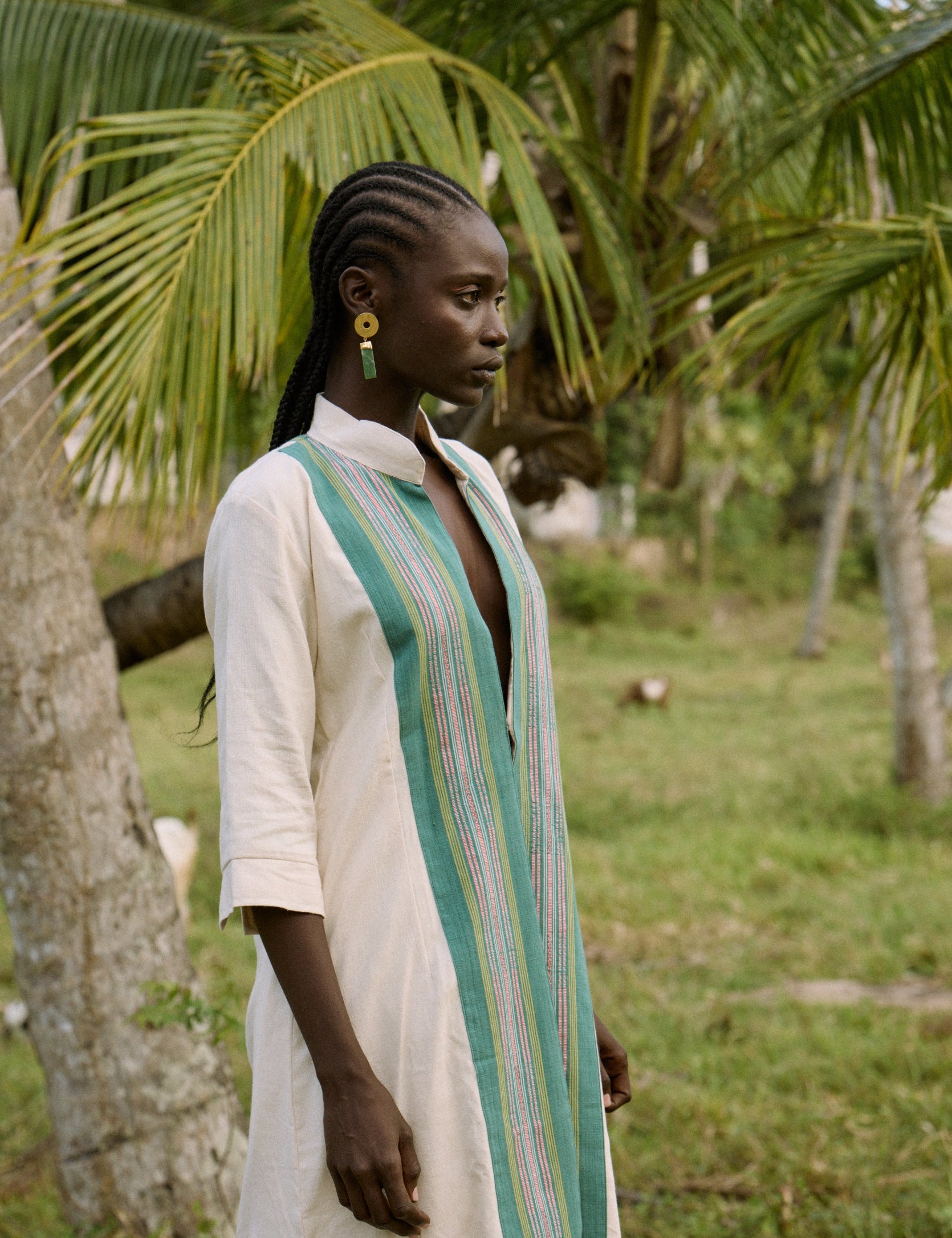 Woman in a long, light-colored dress with a green and white striped scarf standing in a natural setting with trees.