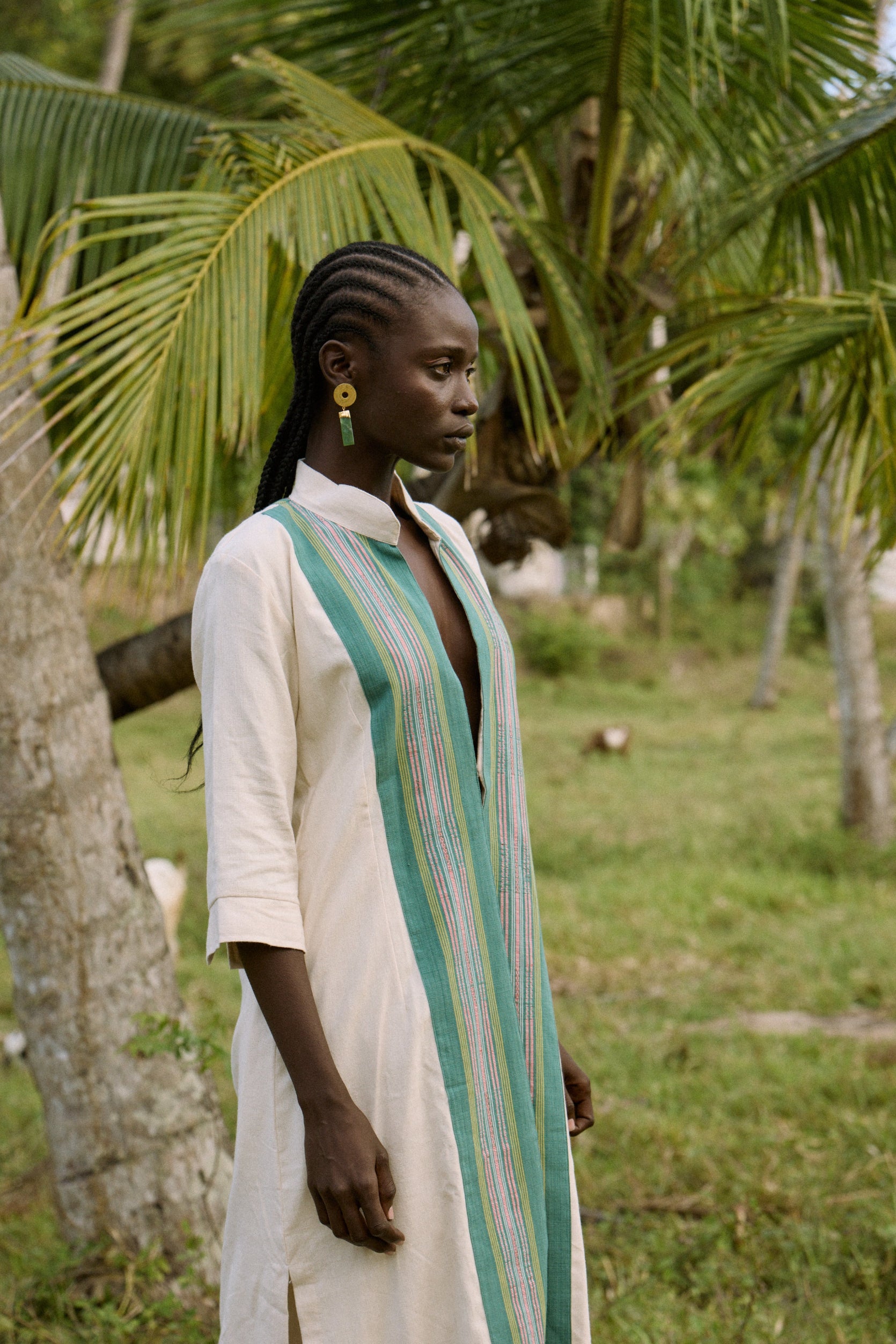 Woman in a long, light-colored dress with a green and white striped scarf standing in a natural setting with trees.